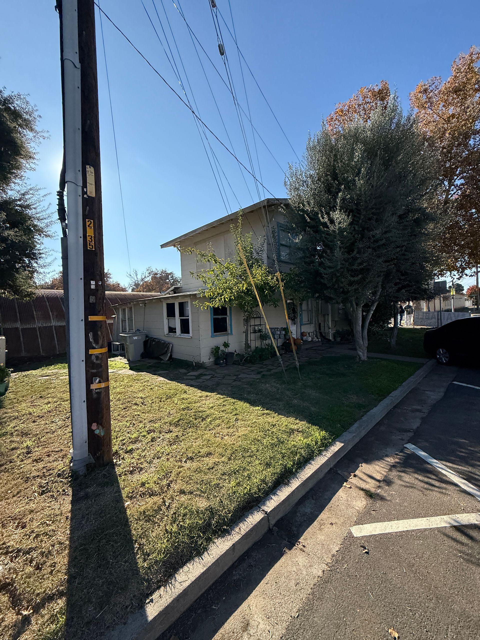 Two-story building with beige siding, trees, and power lines. Green grass in front, under a sunny sky.