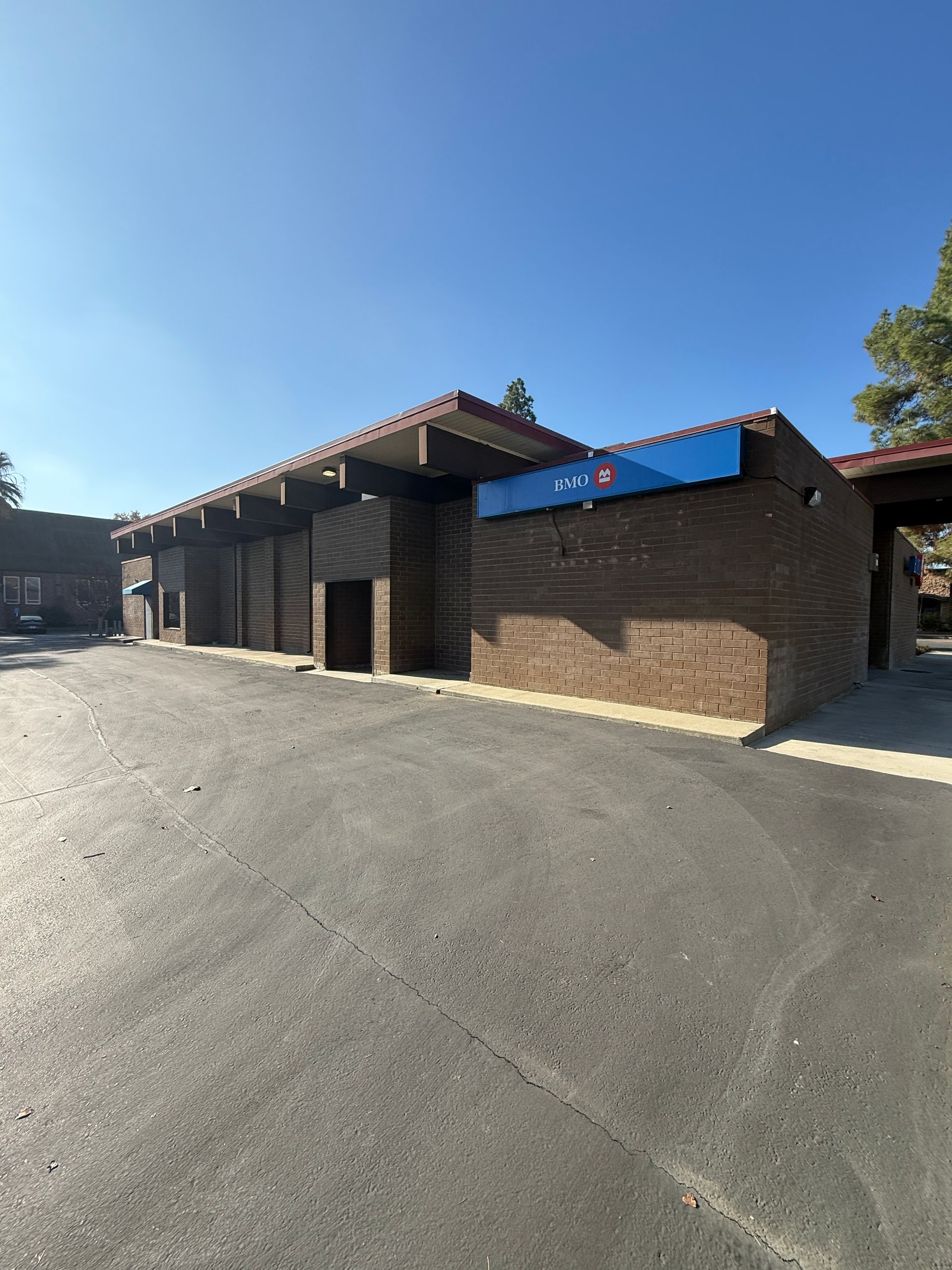 Brick bank building with blue sign, brown roof, and parking area on a sunny day.