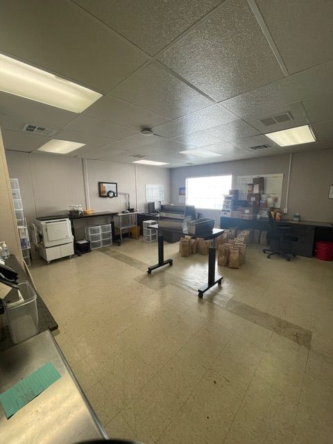 Empty commercial kitchen. Stainless steel counters, appliances, storage. Tables in the center. Neutral tones, fluorescent lighting.