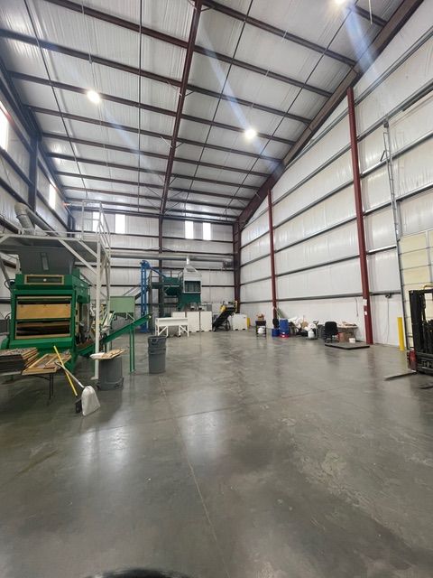 Large warehouse interior with metal frame and concrete floor. Machinery, equipment, and barrels are visible.