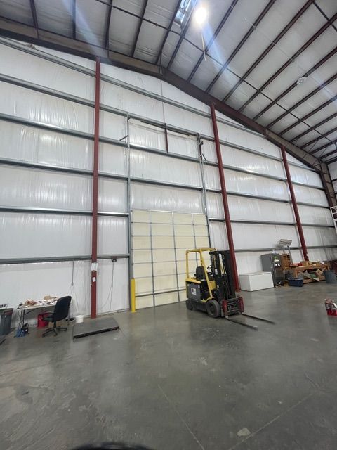 Inside of a warehouse, a forklift is in front of a large roll-up door. Metal and white walls.