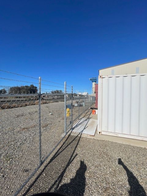 Chain-link fence borders gravel lot next to white industrial building under blue sky.