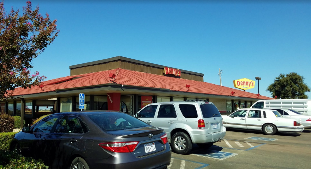 Denny's restaurant exterior with cars in parking lot on a sunny day. Red roof, blue sky, and a sign.