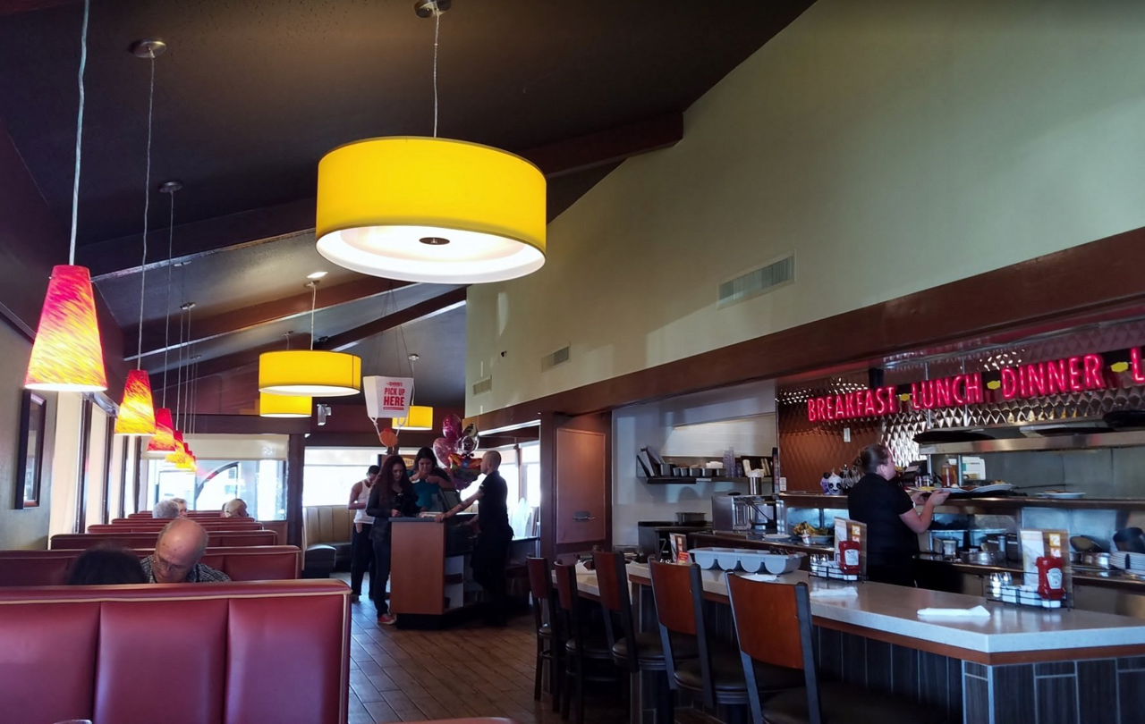 Inside a diner, with red booths and a bar, a waitress serves customers. Yellow and red lamps hang from the ceiling.
