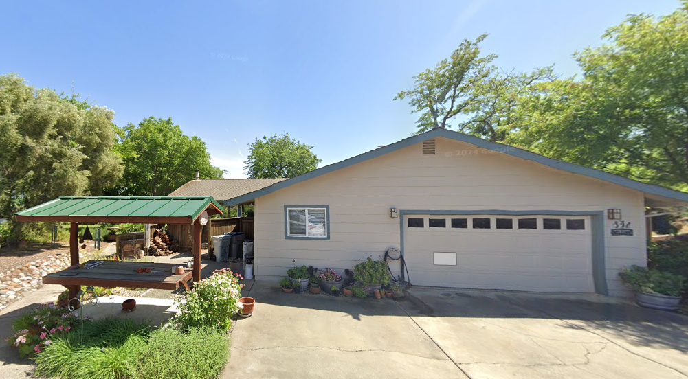 Beige ranch-style house with a two-car garage, a picnic table, and greenery on a sunny day.