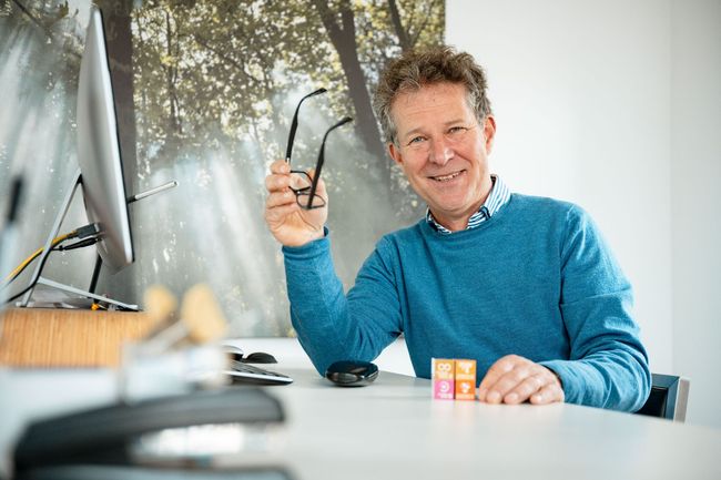 Een man zit aan een bureau met een bril in zijn hand.
