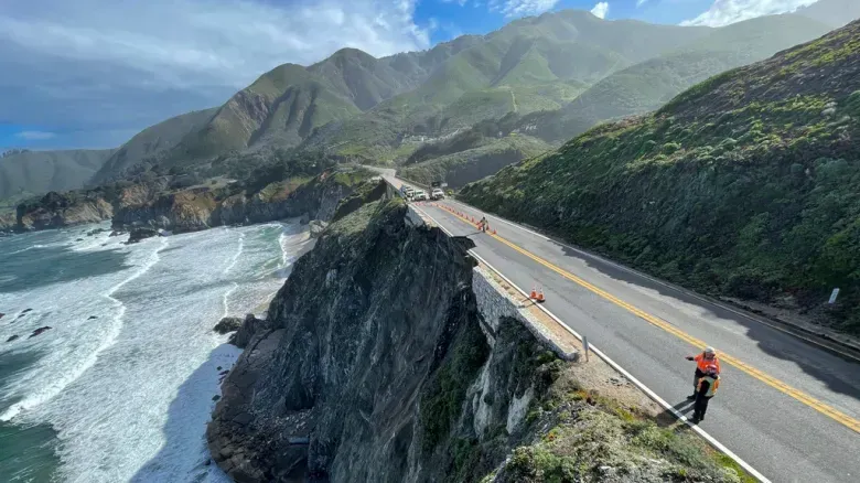 A person is standing on the side of a road next to a cliff overlooking the ocean.