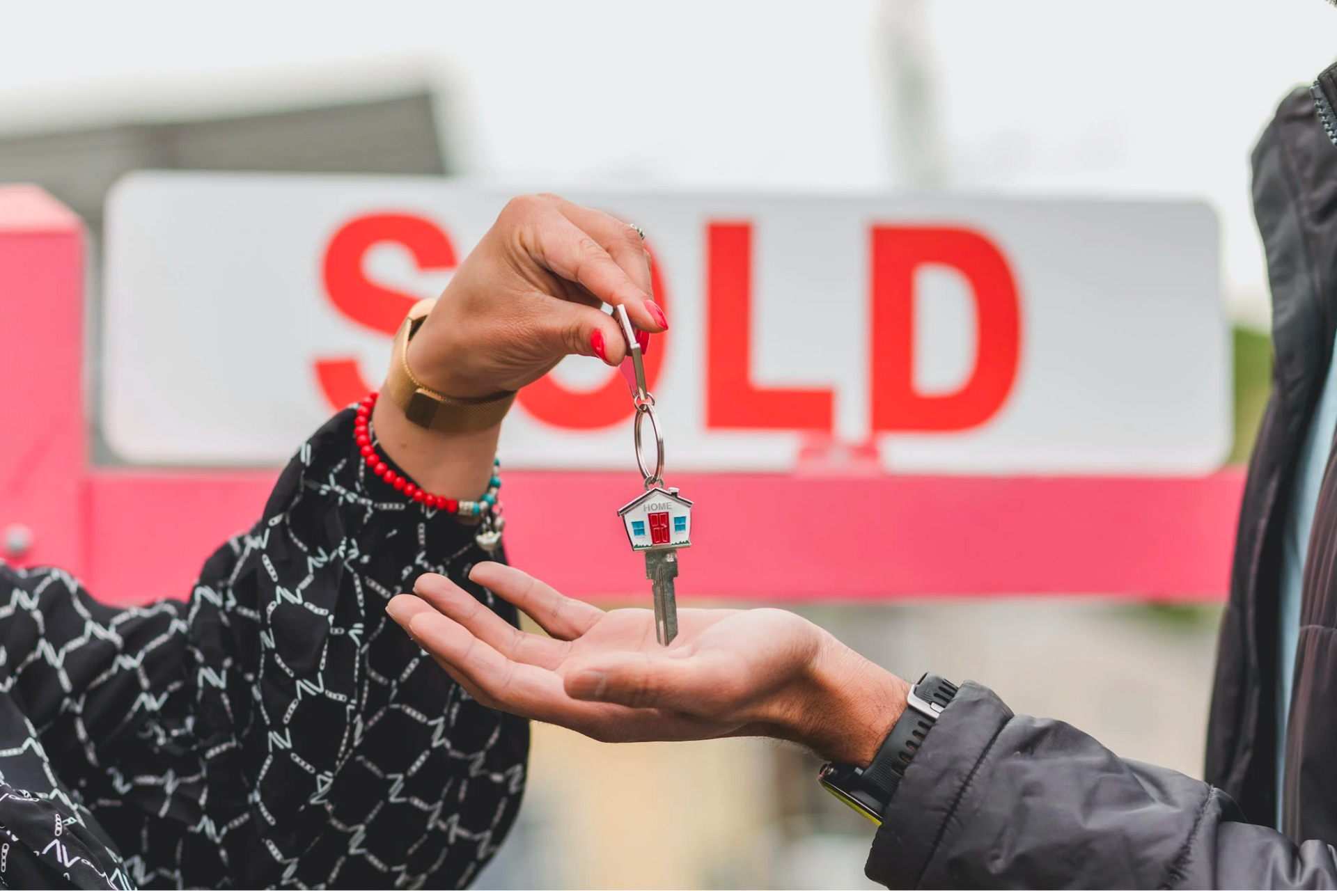 A person is handing a key to another person in front of a sold sign.