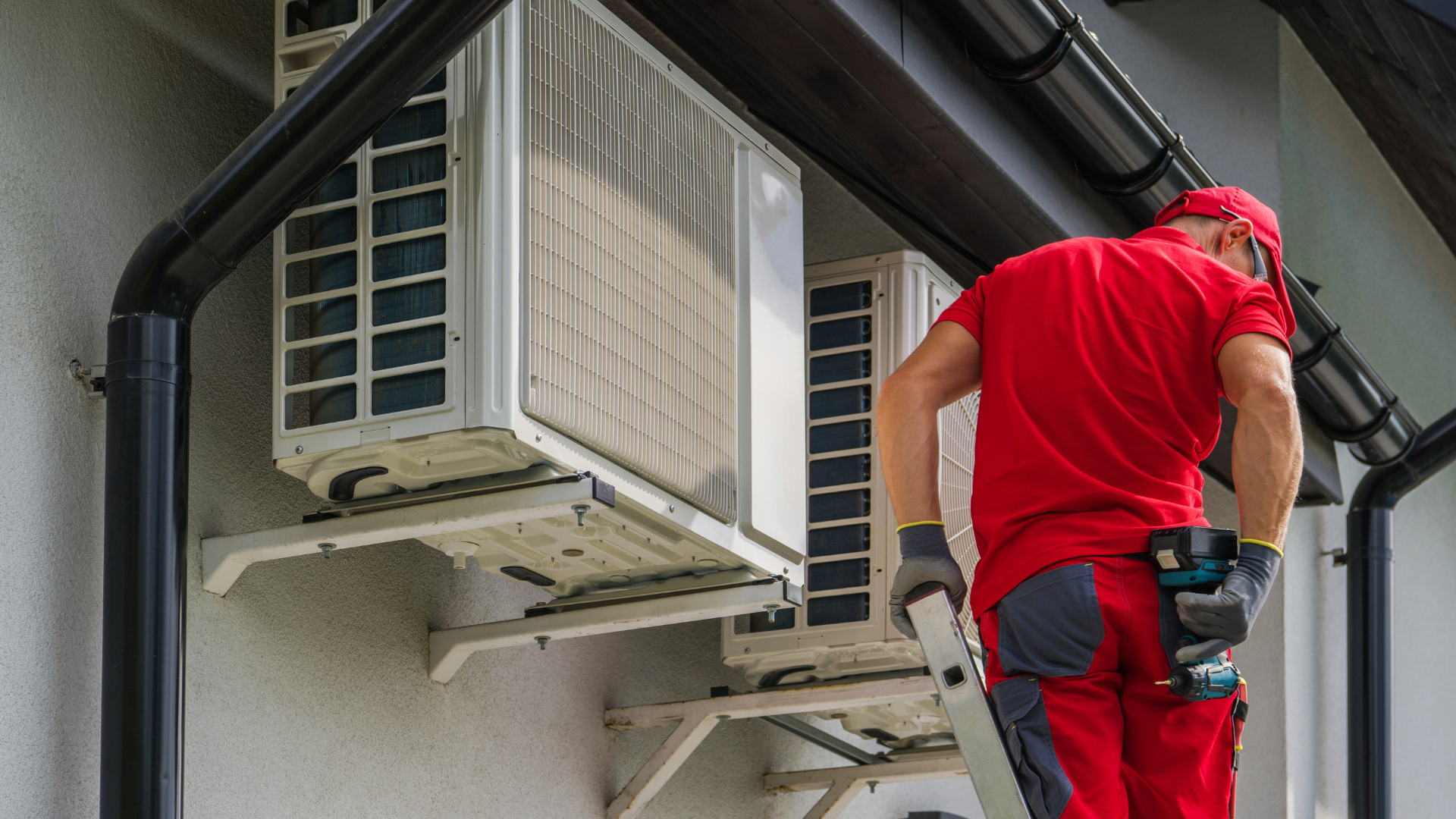 A man is standing on a ladder fixing air conditioners on the side of a building.