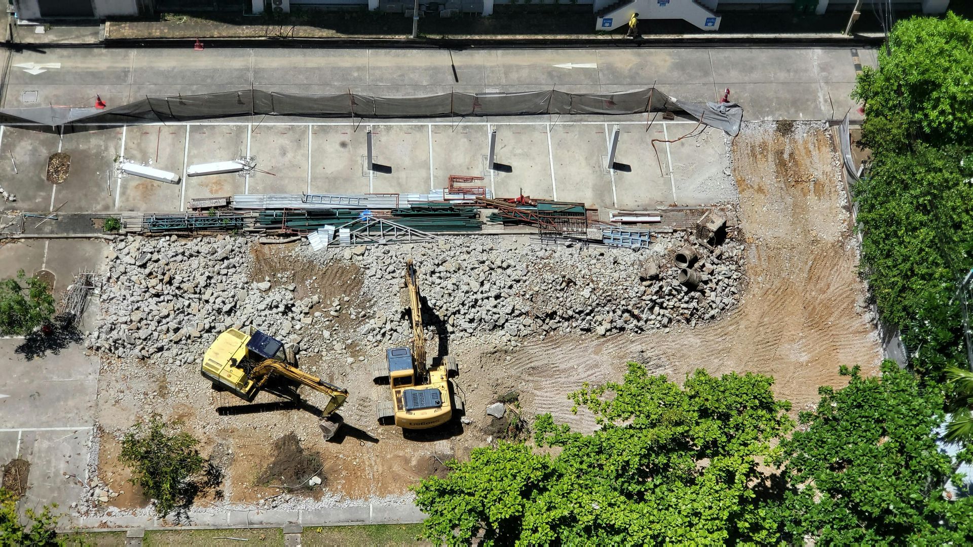 An aerial view shows two yellow construction excavators working on a site with concrete rubble and cleared dirt.