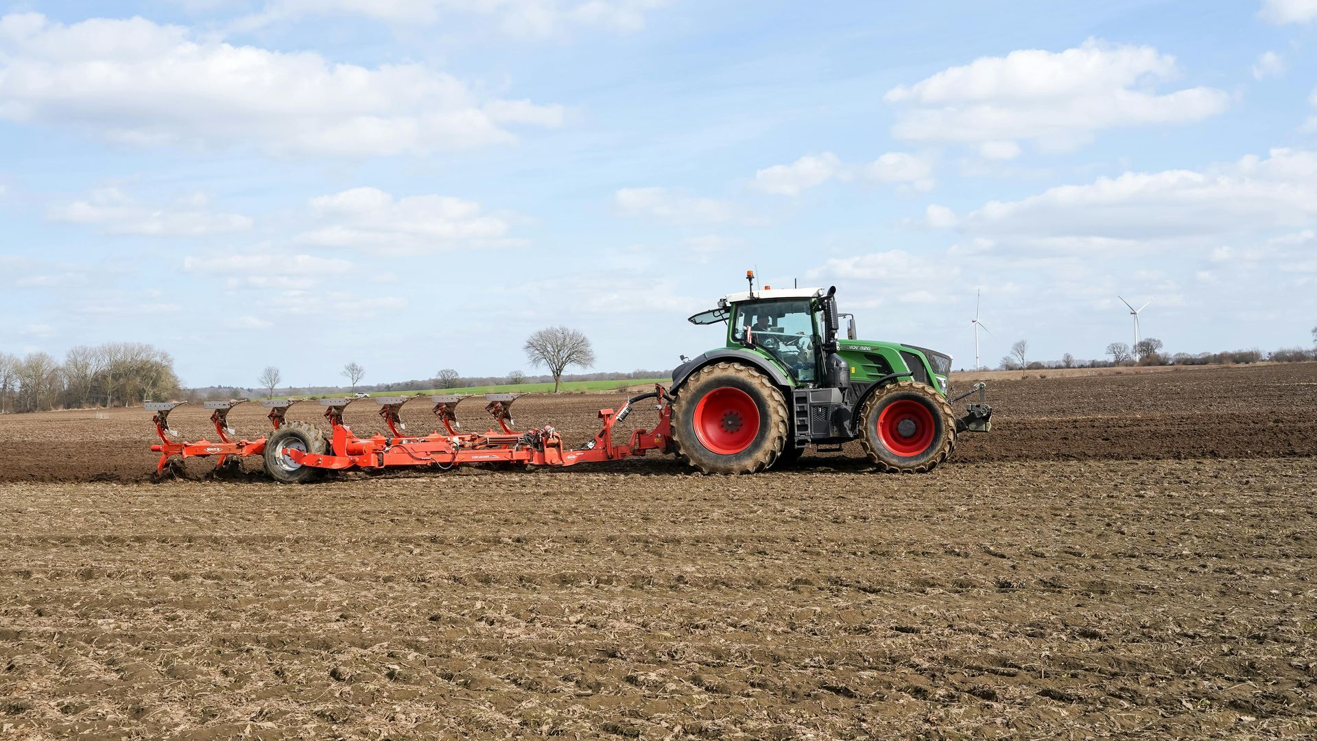 A green Fendt tractor pulls an orange plow through a tilled brown field under a bright, partly cloudy sky.