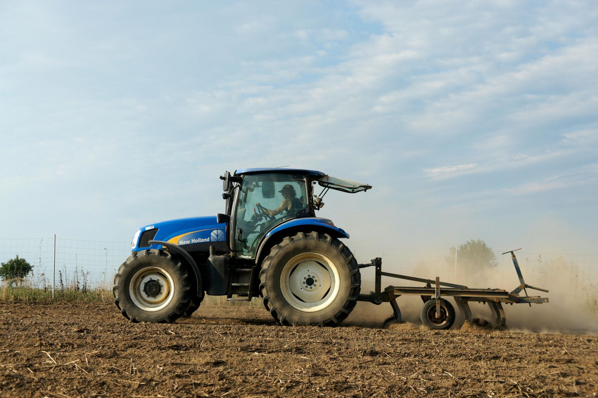 A blue New Holland tractor plows a dry, dusty field under a clear blue sky.