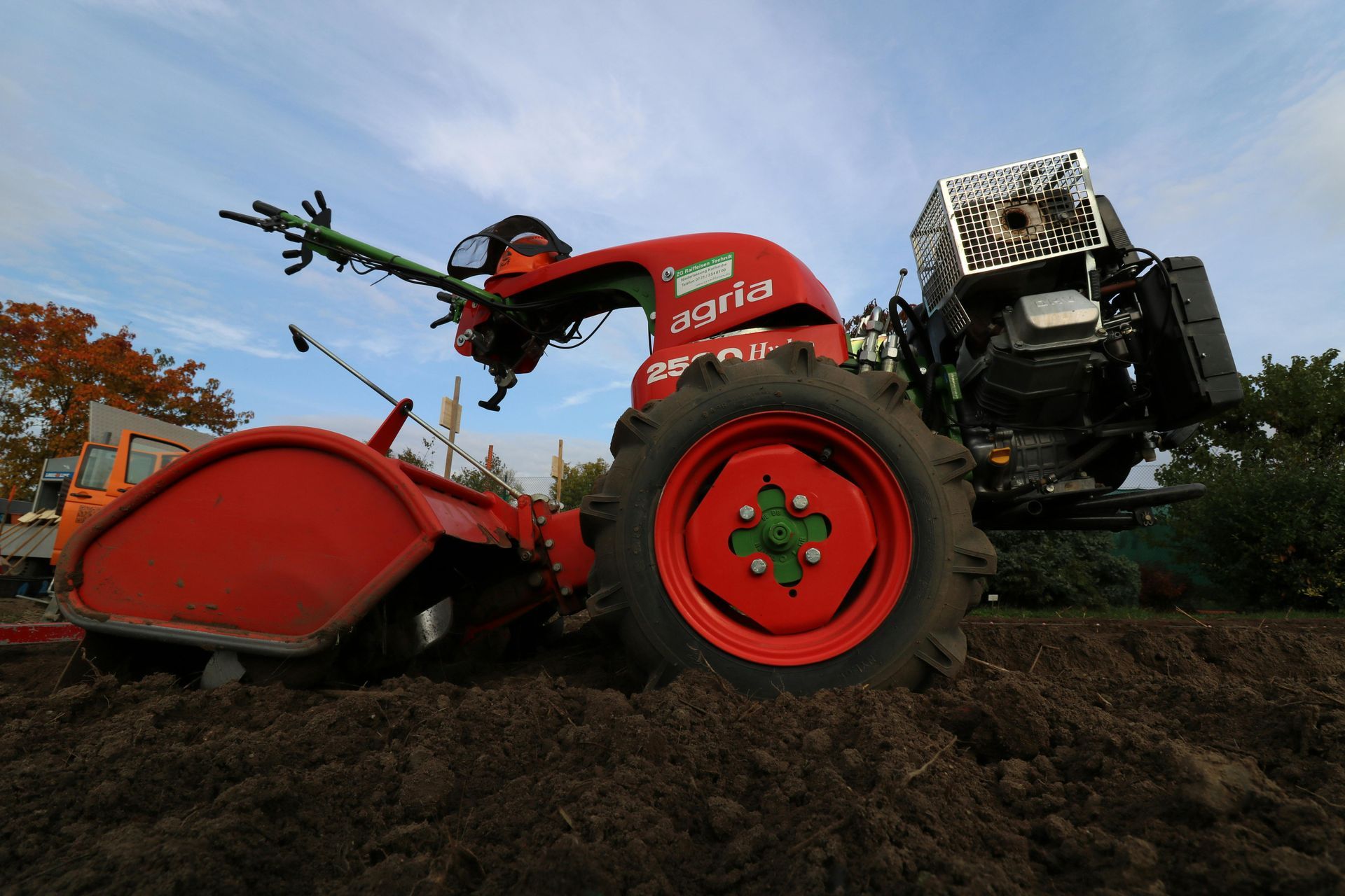 A red Agria walk-behind tractor tilling dark, freshly plowed soil under a blue sky.