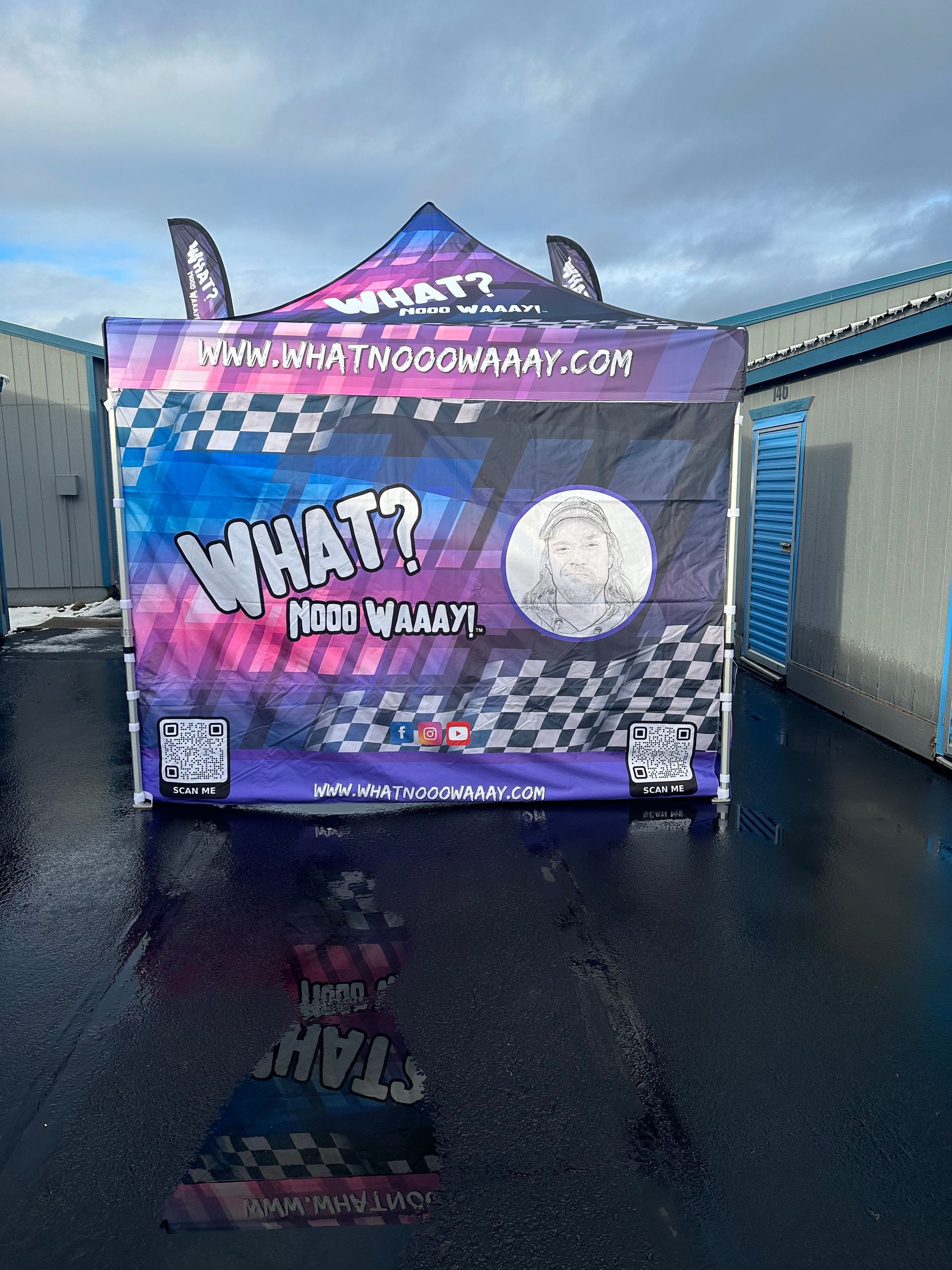 Purple event trailer with “WHAT?” graphics parked on wet pavement under a cloudy sky