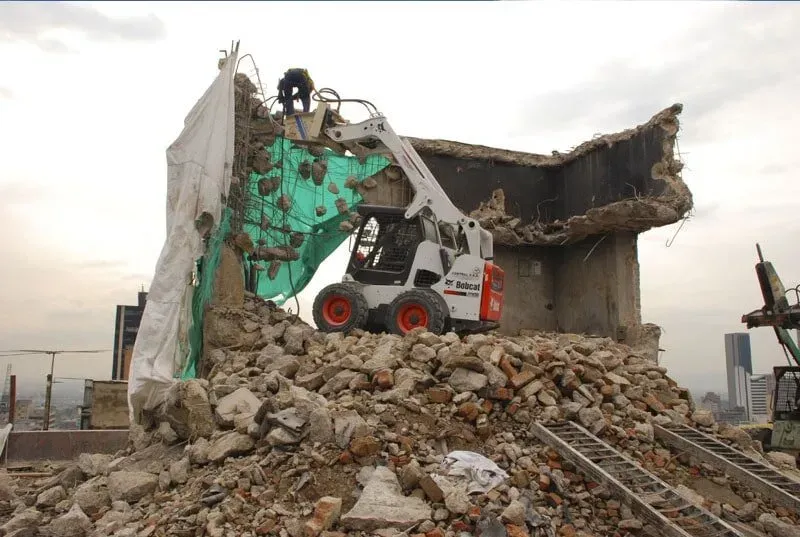 Una excavadora está demoliendo un edificio con un montón de rocas delante.