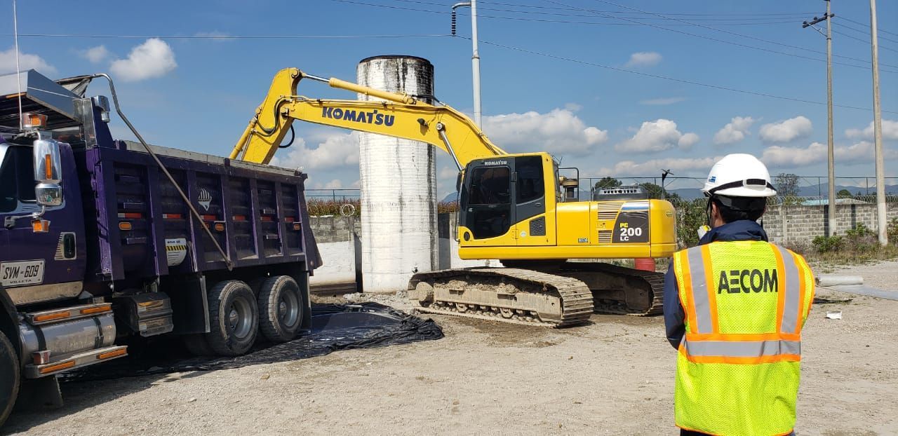 Un hombre con un chaleco amarillo está parado frente a un camión volquete y una excavadora.