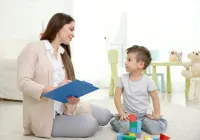Woman with clipboard talking to child playing with blocks on a floor.