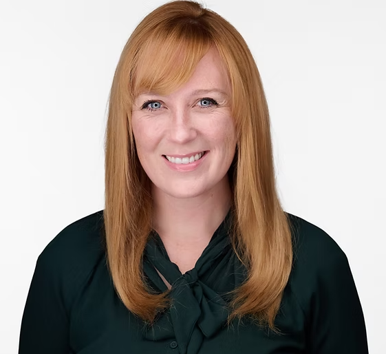 Woman with red hair and bangs, smiling, wearing a dark green blouse, against a white background.