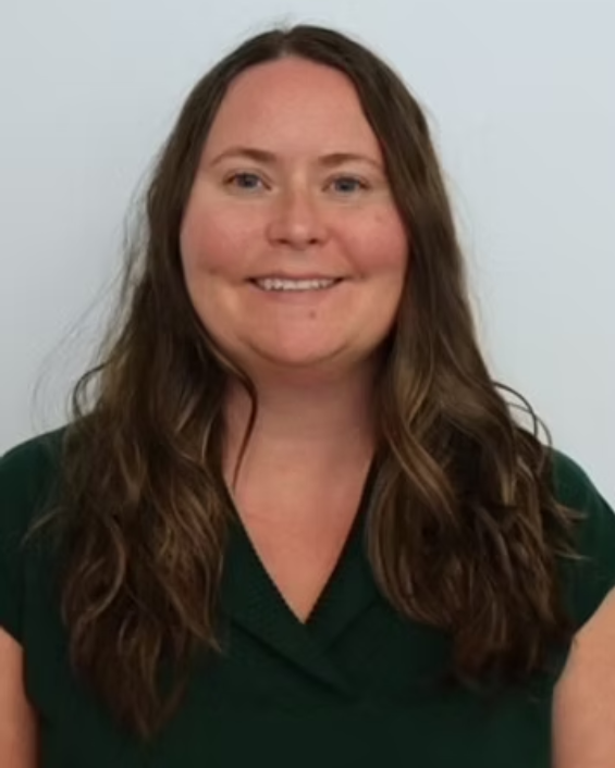 Woman with long brown hair, smiling, wearing a dark green top, against a white backdrop.
