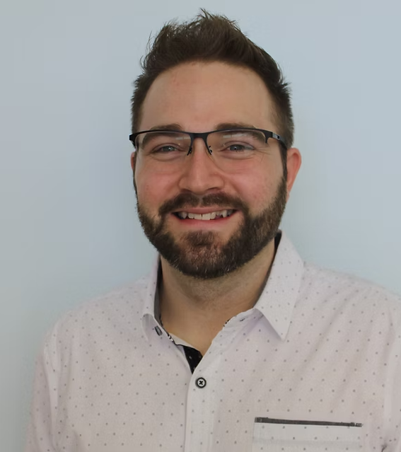 Man with glasses and beard, smiling. Wearing a patterned white shirt, in front of a blue wall.