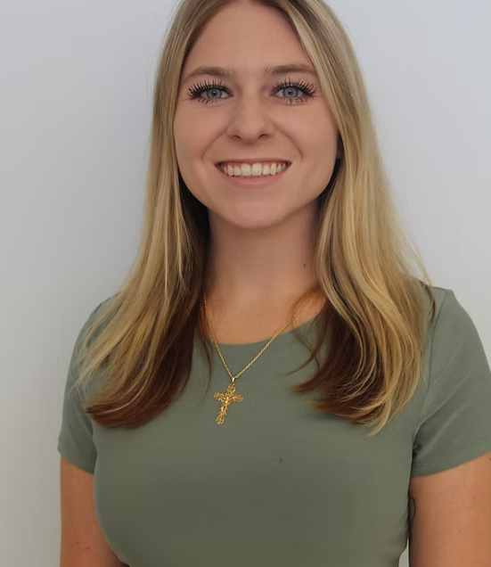 Woman with blonde hair, smiling, wearing a green shirt and gold cross necklace, standing in front of a white wall.
