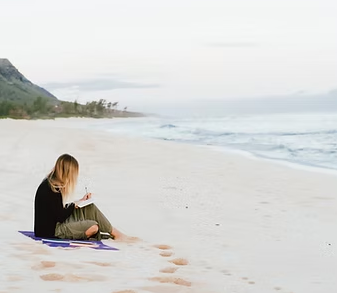 Woman sketching in notebook on a beach. Ocean waves in background. Cloudy sky.