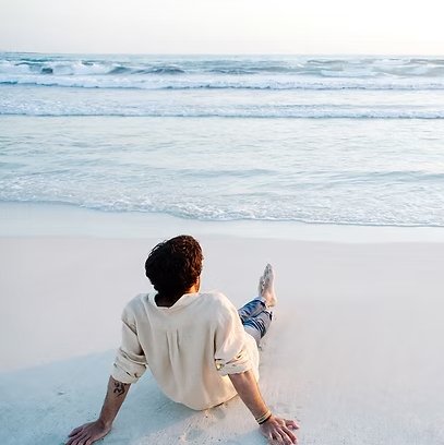 Person sitting on a sandy beach, looking out at the ocean waves.
