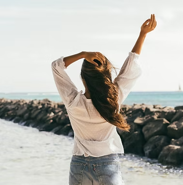Woman on beach raises one arm, touching hair. Wearing white shirt and blue jeans, sunny day.