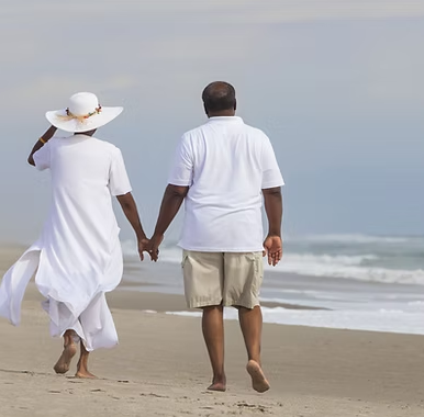 Couple holding hands, walking barefoot on a beach near the ocean. One wears white, the other khakis.