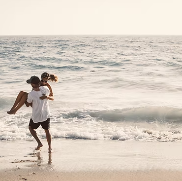 Man carrying a person on his back into the ocean; beach setting with waves and a bright sky.