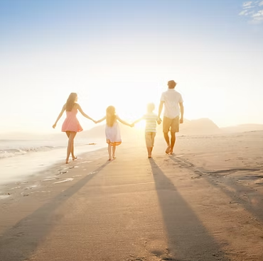Family holding hands walking on a sandy beach towards the bright sun.