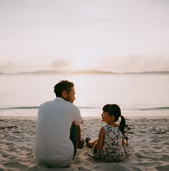 Man and child sitting on a beach, looking at each other, ocean and sunset in background.