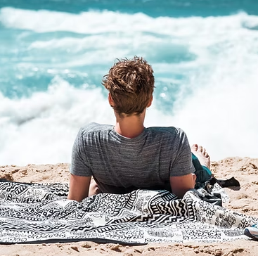 Person lying on a beach towel, facing the ocean. Blue water, white waves, sunny day.