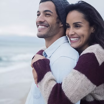 Couple hugging on beach, smiling at something out of frame. She's wearing a sweater, he has a beanie.