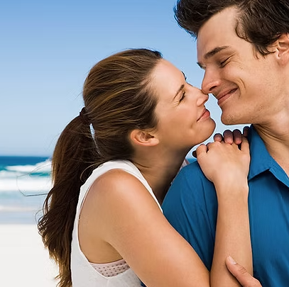 Woman gently touches man's nose; they smile, standing near a beach.