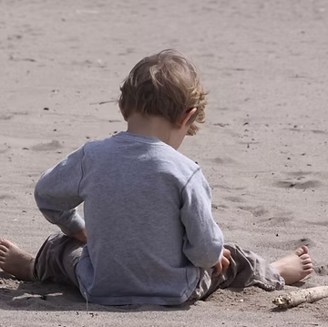 Child sitting on sandy beach, looking down. Gray shirt, brown pants. Bare feet.