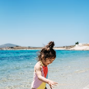 Child in colorful swimsuit at the beach, reaching into the water on a sunny day.