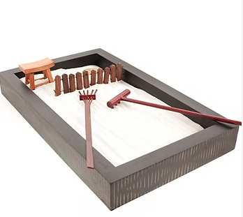 Miniature zen garden with dark wood frame, sand, wooden rake, and small fence.