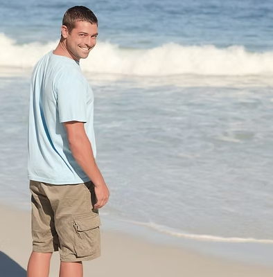 Man on beach, wearing light blue shirt and khaki shorts, smiling, ocean in background.