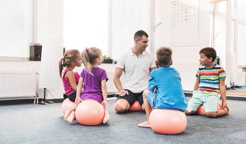 Children and instructor sit on exercise balls in a room, conversing.