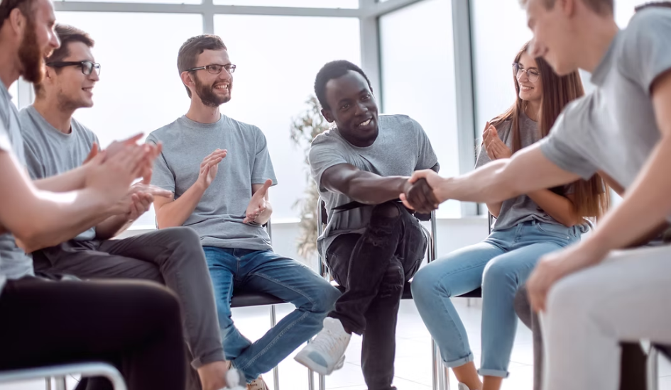 People in a circle, smiling and clapping, two shaking hands, white room setting.