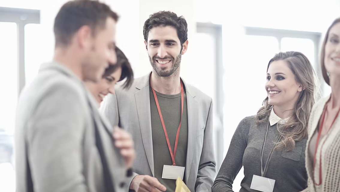 Group of people smiling, wearing lanyards, interacting in a well-lit indoor setting.