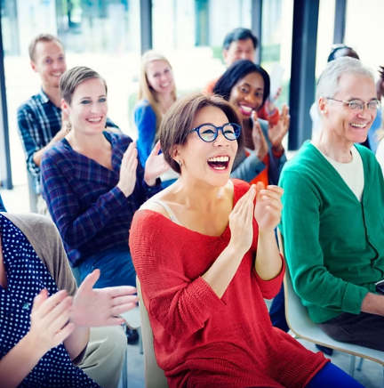 Audience clapping, laughing, seated in a brightly lit room. Woman in red sweater laughs, center focus.