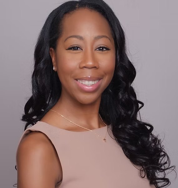 Woman with blonde hair, smiling, wearing a green shirt and gold cross necklace, standing in front of a white wall.