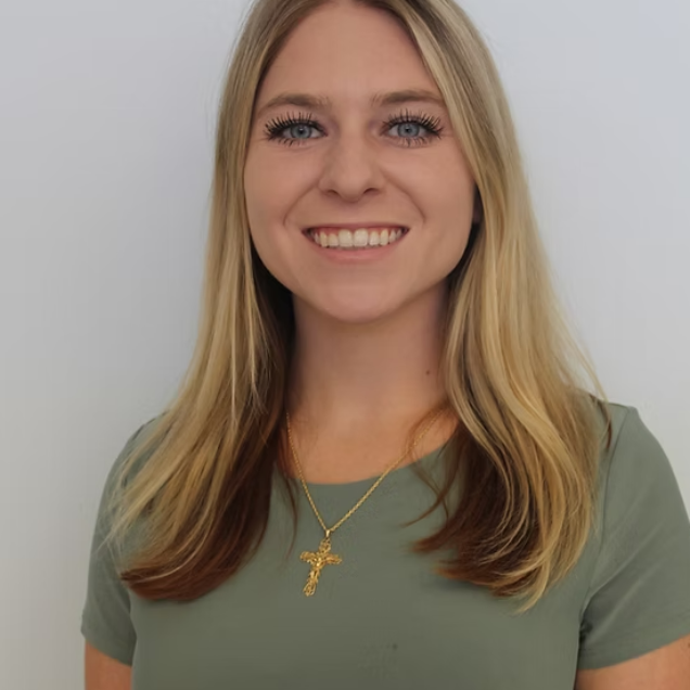 Woman with blonde hair, smiling, wearing a green shirt and gold cross necklace, standing in front of a white wall.