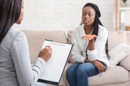 Woman gesturing while speaking to another woman taking notes on a clipboard in a counseling session.