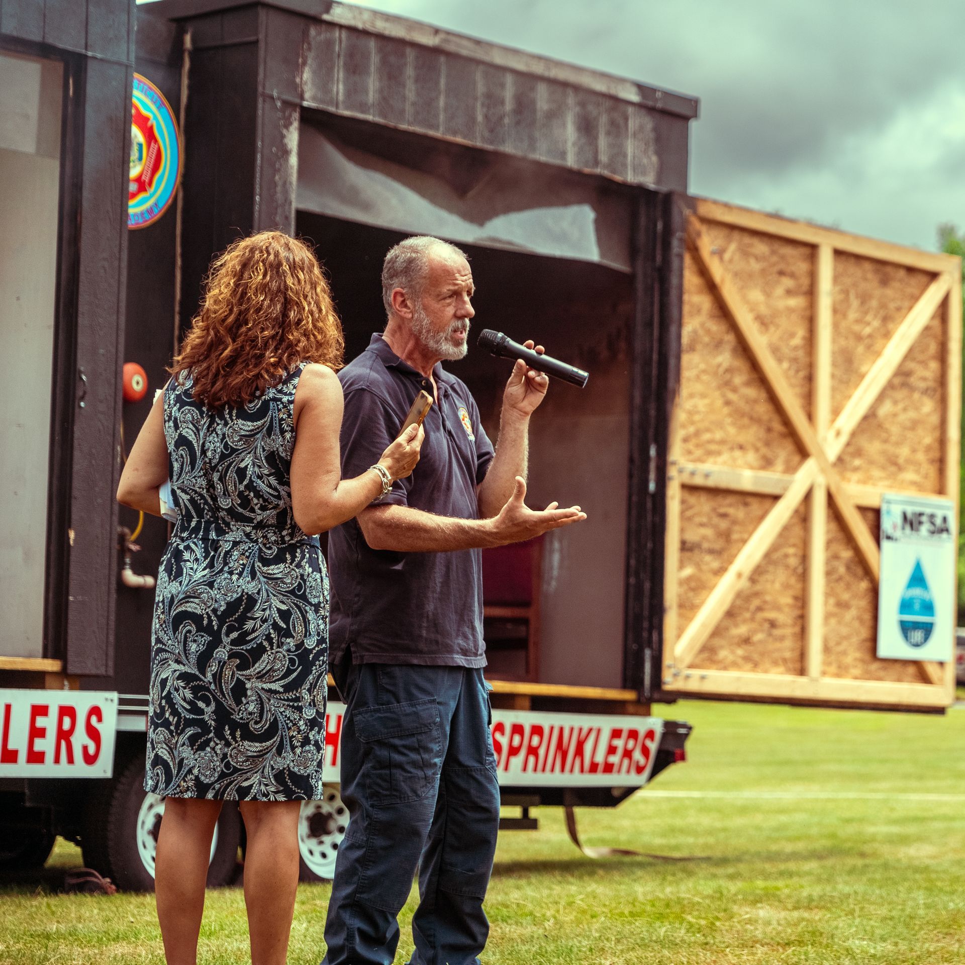 A man speaking into a microphone in front of a trailer that says sprinklers