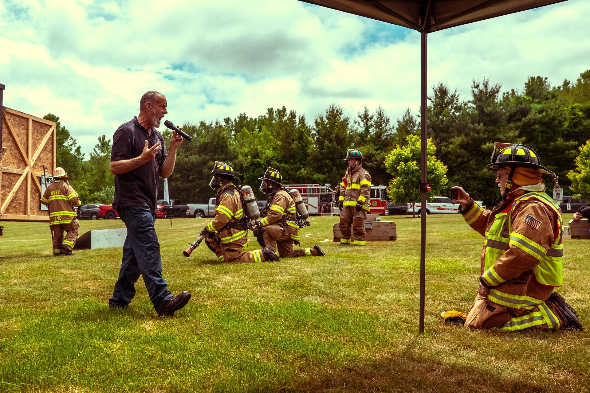 A man is talking into a microphone while firefighters are kneeling down in a field.