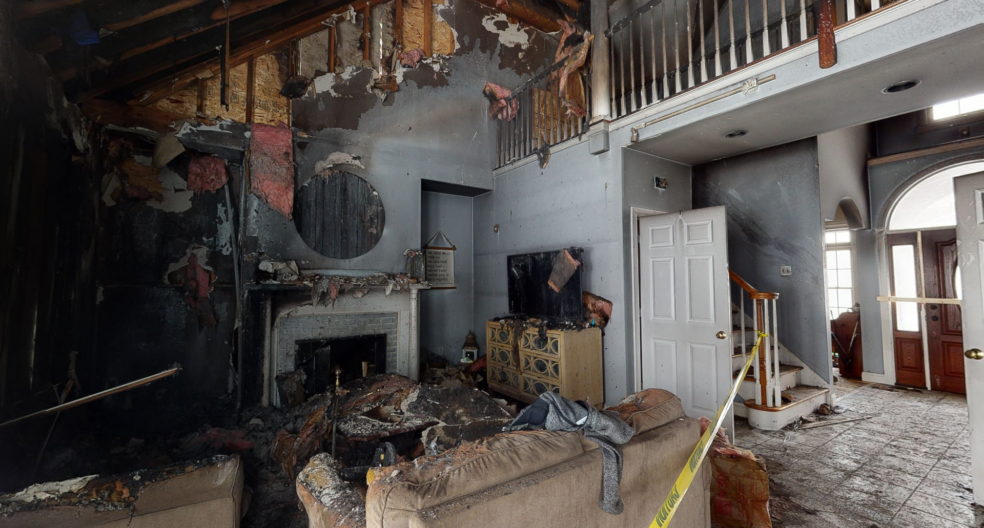 Interior of a fire-damaged home. Charred walls, debris, and burnt furniture are visible; yellow caution tape is present.