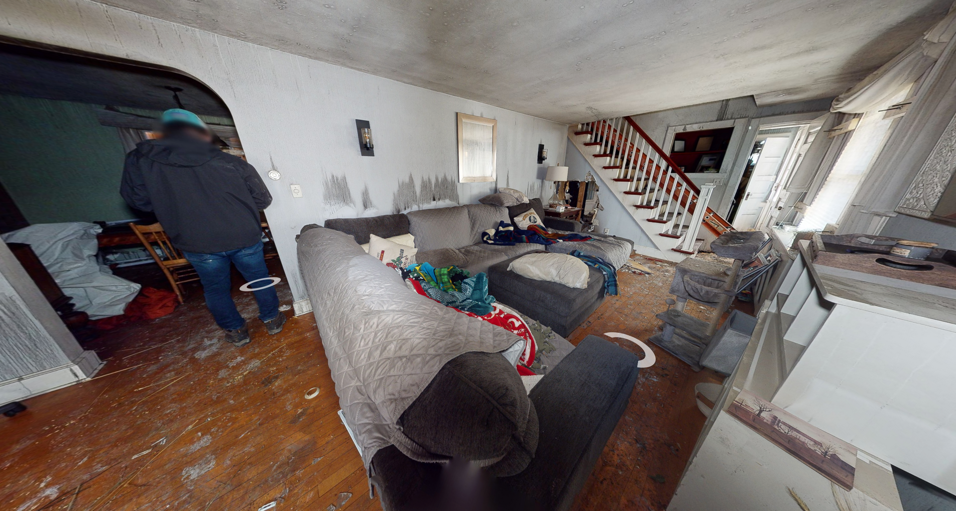 Man in a dark jacket in a messy living room with a damaged couch and staircase.